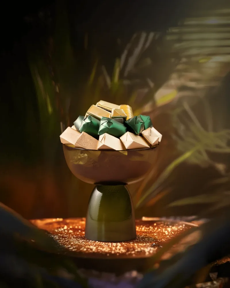 Coupe de verre vert garnie de chocolats vert et or dans un décor de palmes