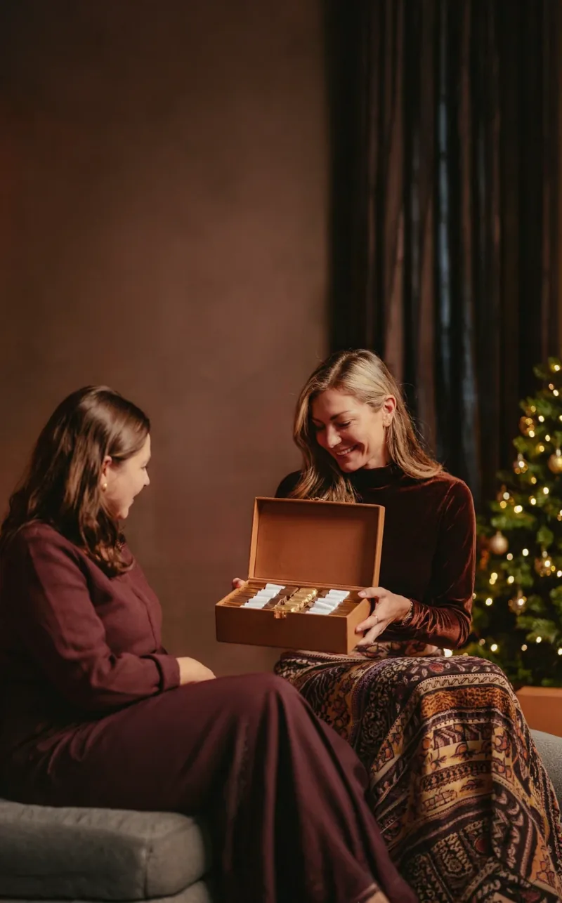 Une femme ouvre un coffret NAYA en cuir camel offert par une amie, près d'un sapin illuminé